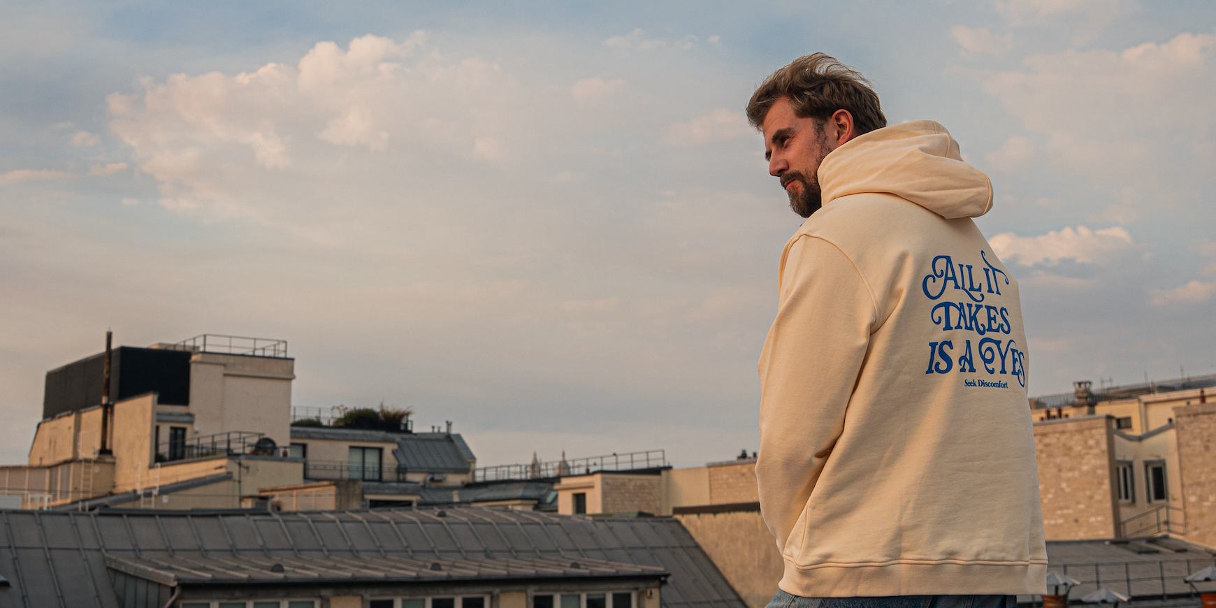 Thomas Brag wearing a cream-colored hoodie with blue text on the back stands on a rooftop overlooking city buildings under a cloudy sky.